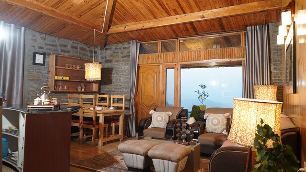 A cozy living room with wooden furniture, a stone wall, and a large window. - The Whispering Pines (Dhari)