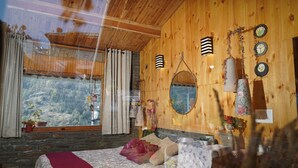 A wooden cabin room with a bed, a window with curtains, a mirror, and wall decorations. - The Whispering Pines (Dhari)