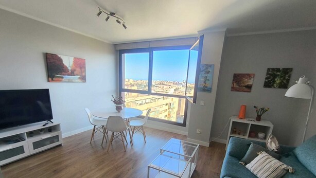 A modern living room with a large window, a flat-screen TV, a dining table, and a blue sofa. - Beautiful apartment in Teatinos Málaga (Málaga)