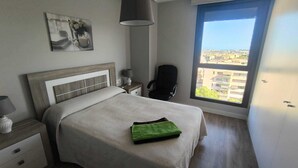 A modern bedroom with a large bed, a nightstand, a chair, a window with a city view, and a hanging lamp. - Beautiful apartment in Teatinos Málaga (Málaga)