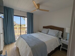 Un dormitori amb llit, una tauleta de nit, una làmpada, un ventilador de sostre i vista a l'exterior.