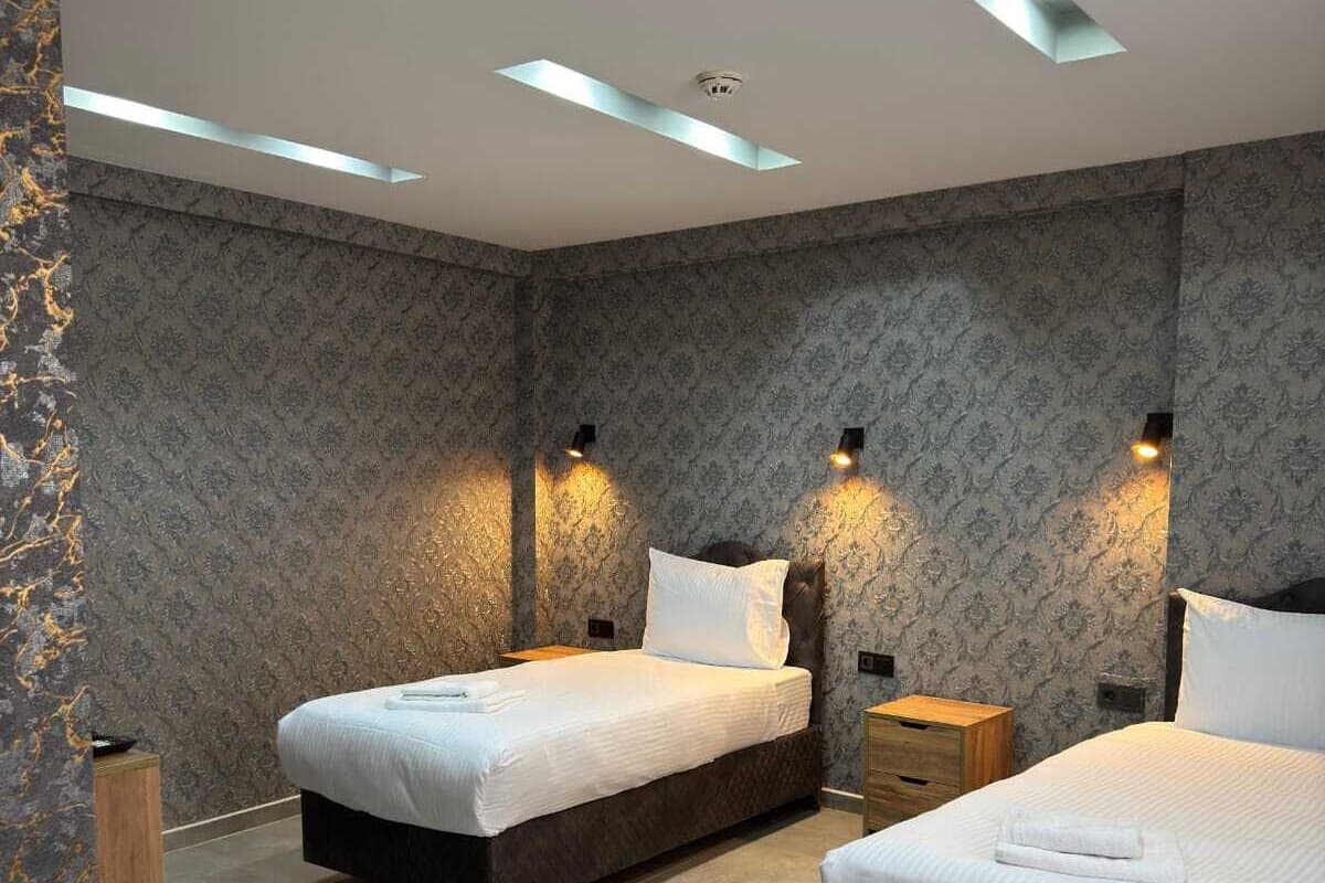 A modern bedroom with two beds, patterned wallpaper, recessed ceiling lights, and a small bedside table with a lamp.