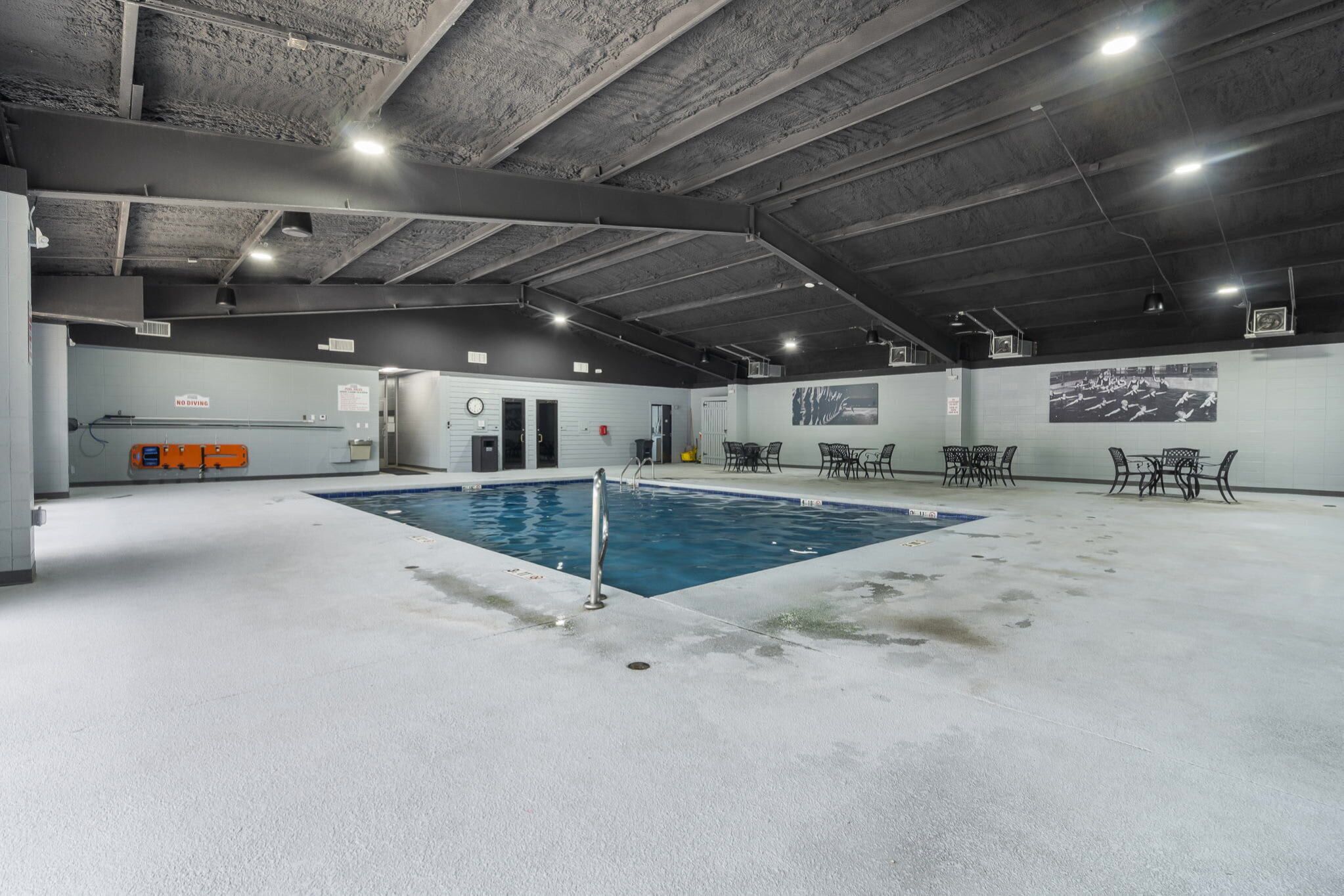 An indoor swimming pool with a concrete floor, surrounded by tables and chairs, and a high ceiling with visible beams.