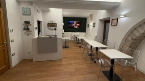 A modern dining area with a long table and chairs, a small reception desk, and a TV screen displaying a cityscape. - Hotel Lieto Soggiorno (Assisi)