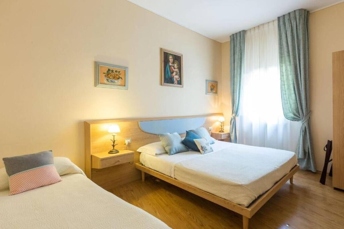 A hotel room with a large bed, a smaller bed, wooden headboard, two bedside tables with lamps, and a window with curtains.