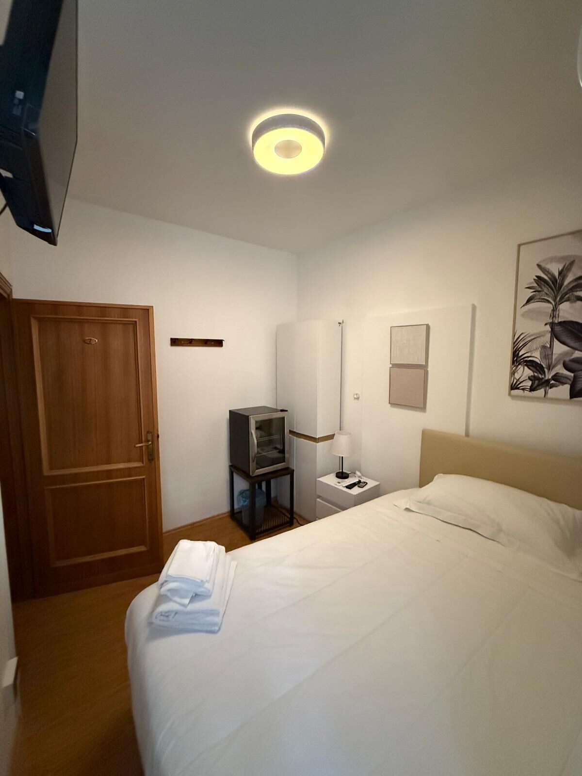A hotel room with a bed, a television on a stand, a nightstand with a lamp, and a door.