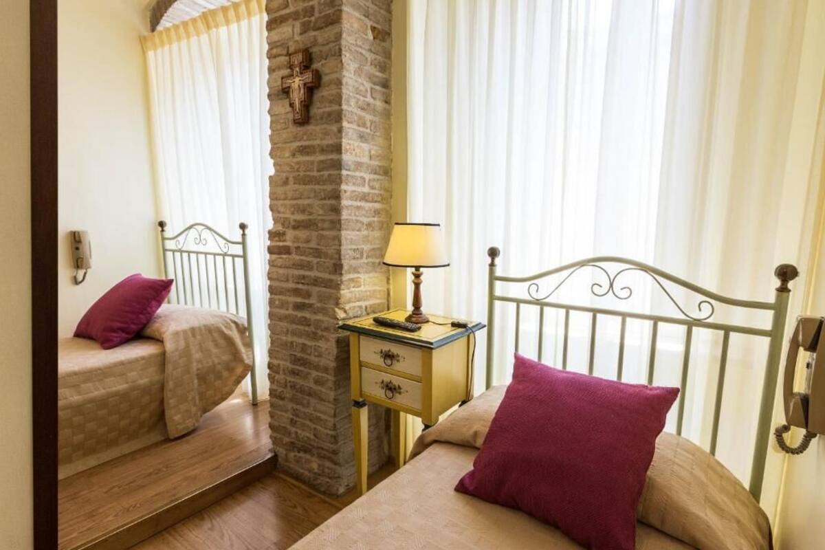 A room with two beds, a brick wall, a small bedside table with a lamp, and a cross on the wall.