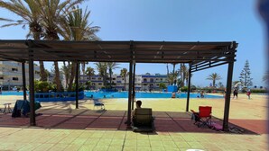 A covered outdoor area with a pool, palm trees, and a building in the background.