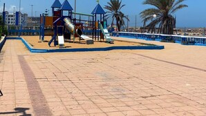 A playground with slides and climbing structures, surrounded by palm trees and a clear blue sky.