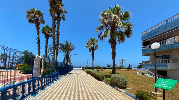 A paved walkway lined with palm trees and a blue fence, leading to a building with balconies and a sign in a foreign language.