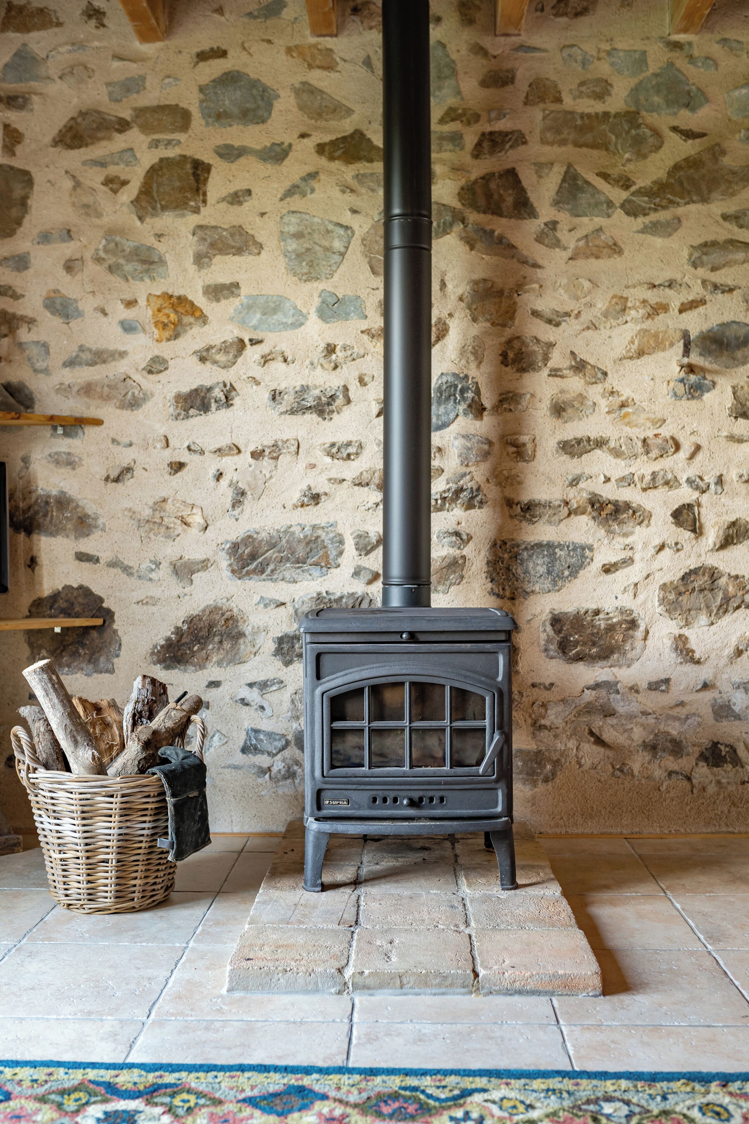 A stone wall with a black wood-burning stove, a wicker basket with firewood, and a tiled floor.