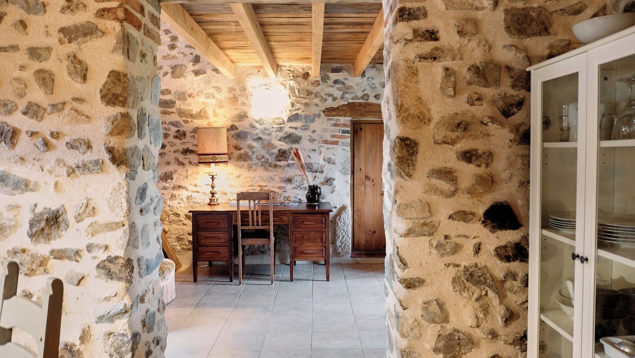 A stone-walled room with a wooden desk, chair, and a cabinet with glass doors.