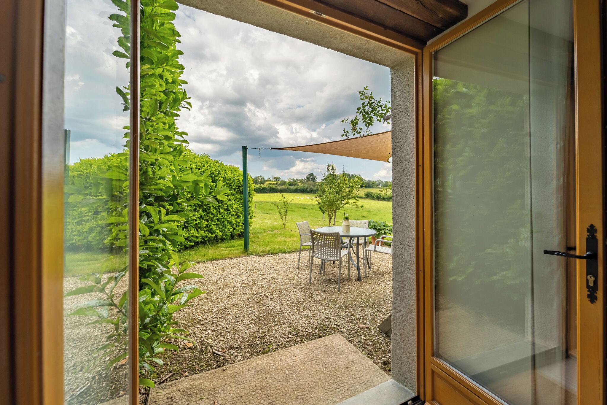 A glass door leading to a patio with a table and chairs, surrounded by greenery and a gravel path.