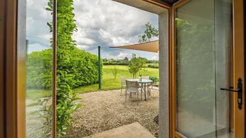 A glass door leading to a patio with a table and chairs, surrounded by greenery and a gravel path.