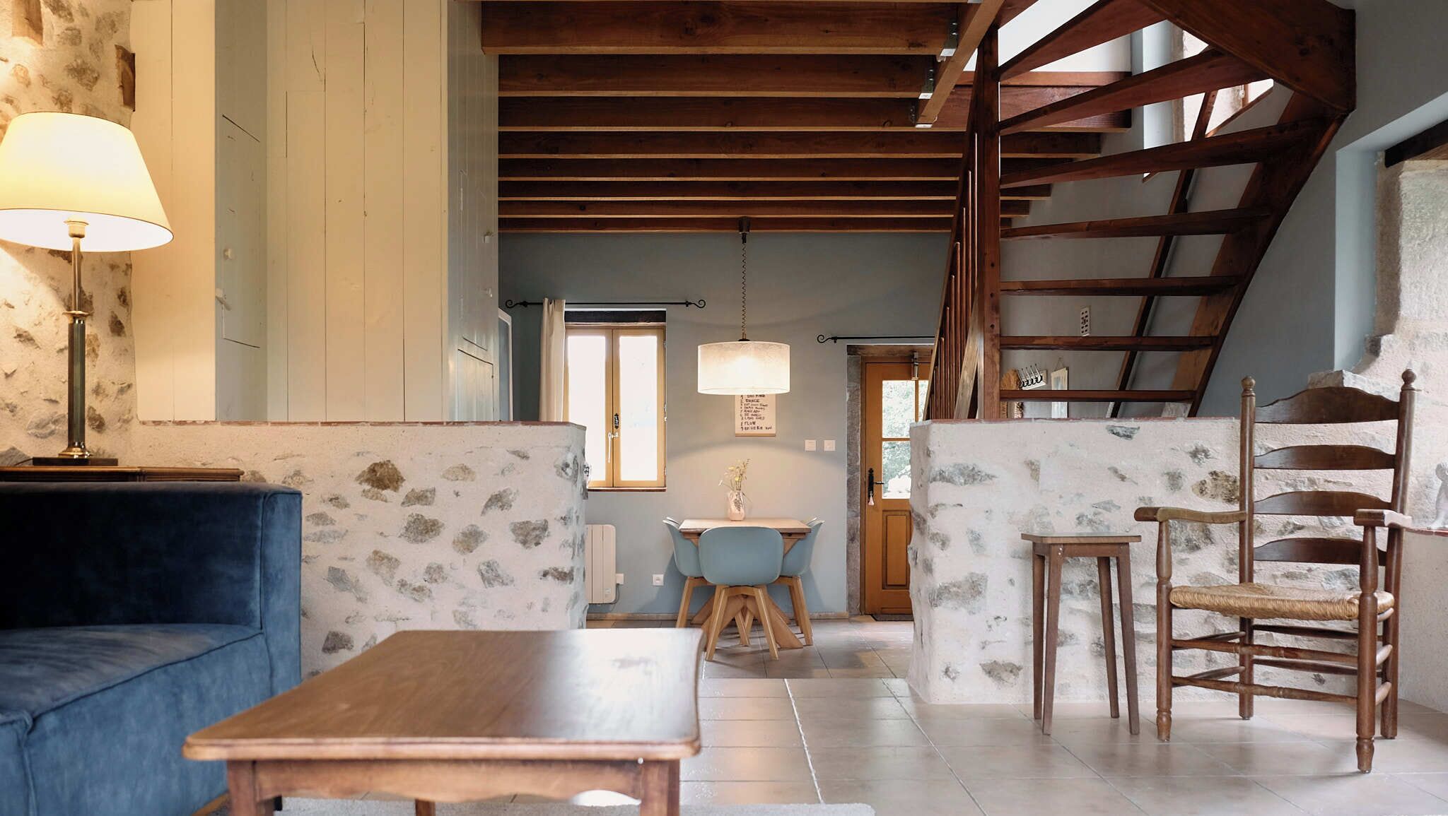 A rustic interior with exposed wooden beams, a stone wall, a blue sofa, a wooden table, and a dining area with a small table and chairs.