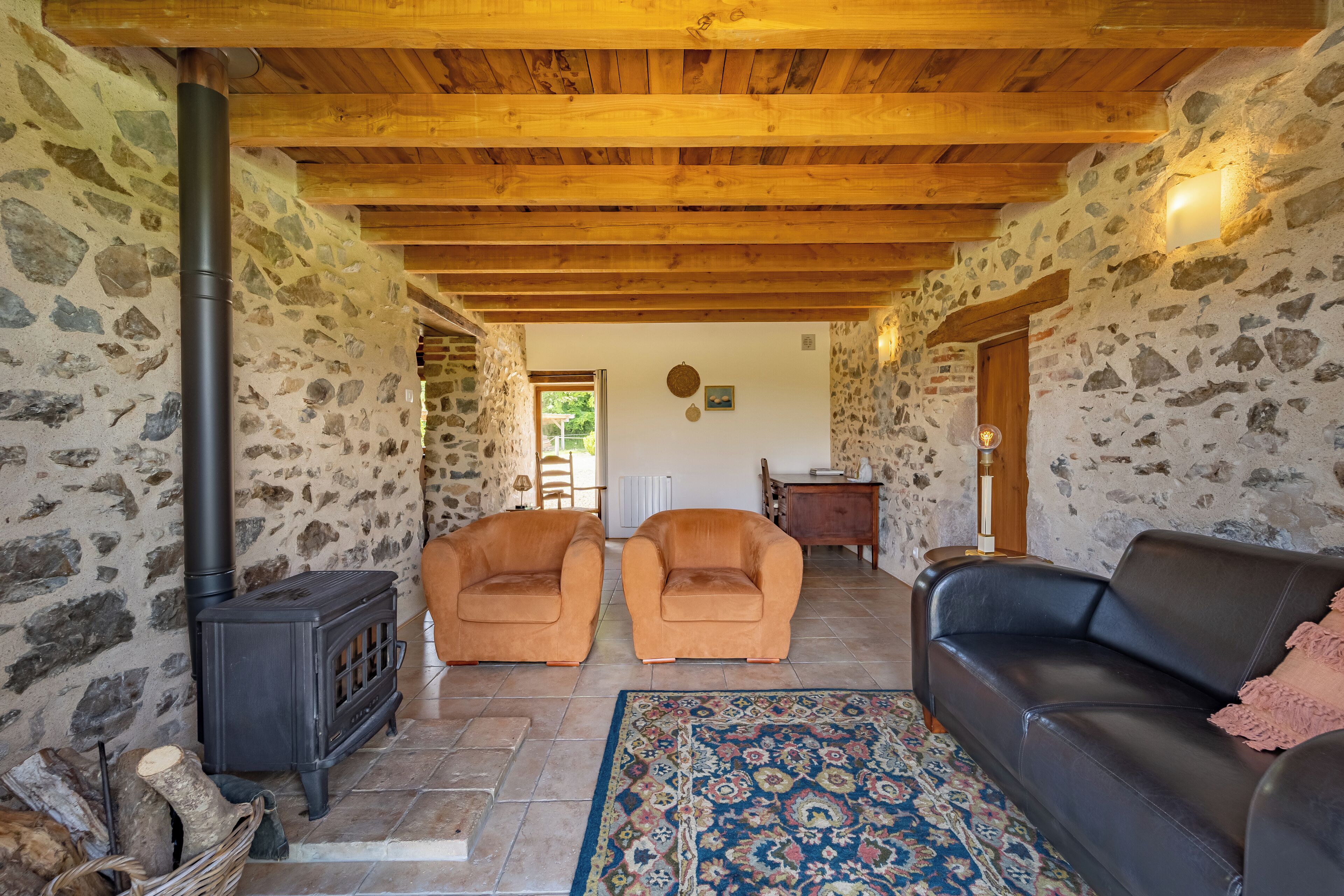 Ein Zimmer mit Steinwänden und Holzbalkendecke, einem schwarzen Sofa, zwei Sesseln, einem gemusterten Teppich und einem Kaminofen.