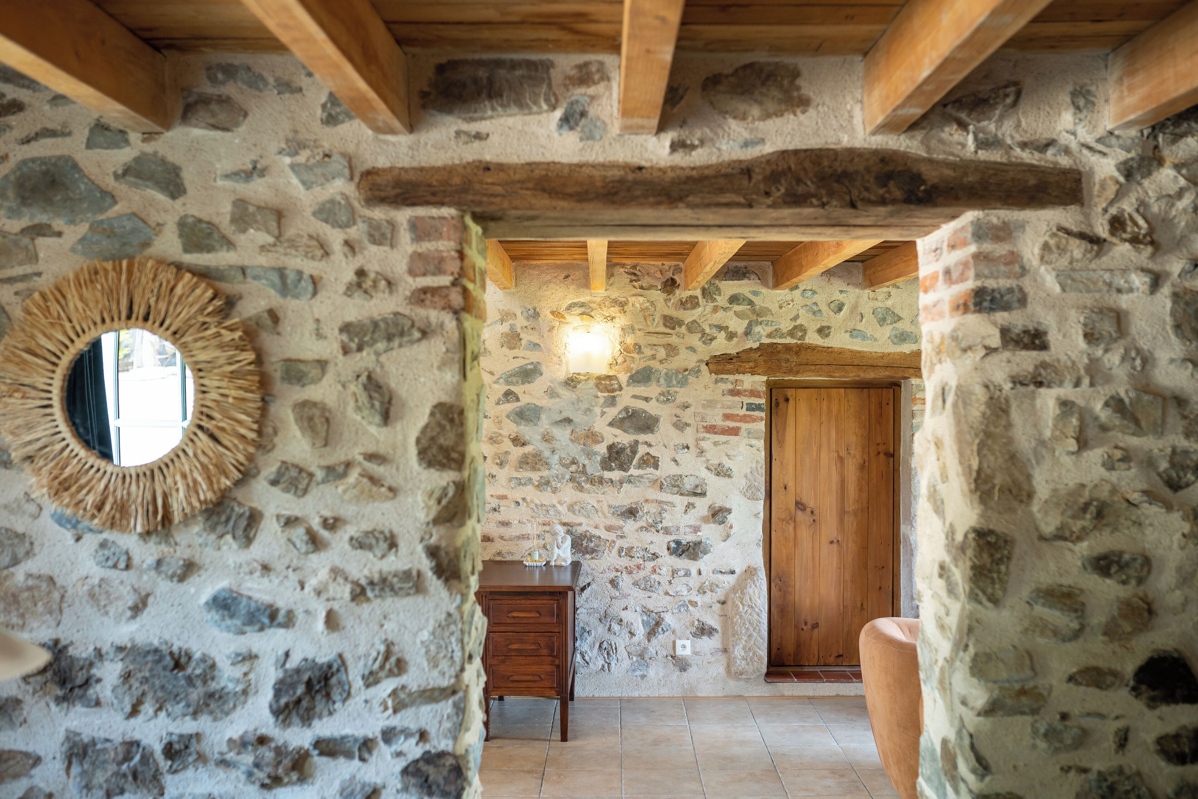 Una habitación rústica con paredes de piedra, techo de madera, un espejo redondo, un aparador de madera y una puerta.