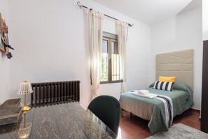 Une chambre à coucher avec un lit, une chaise, une table, un radiateur et une fenêtre avec des rideaux.