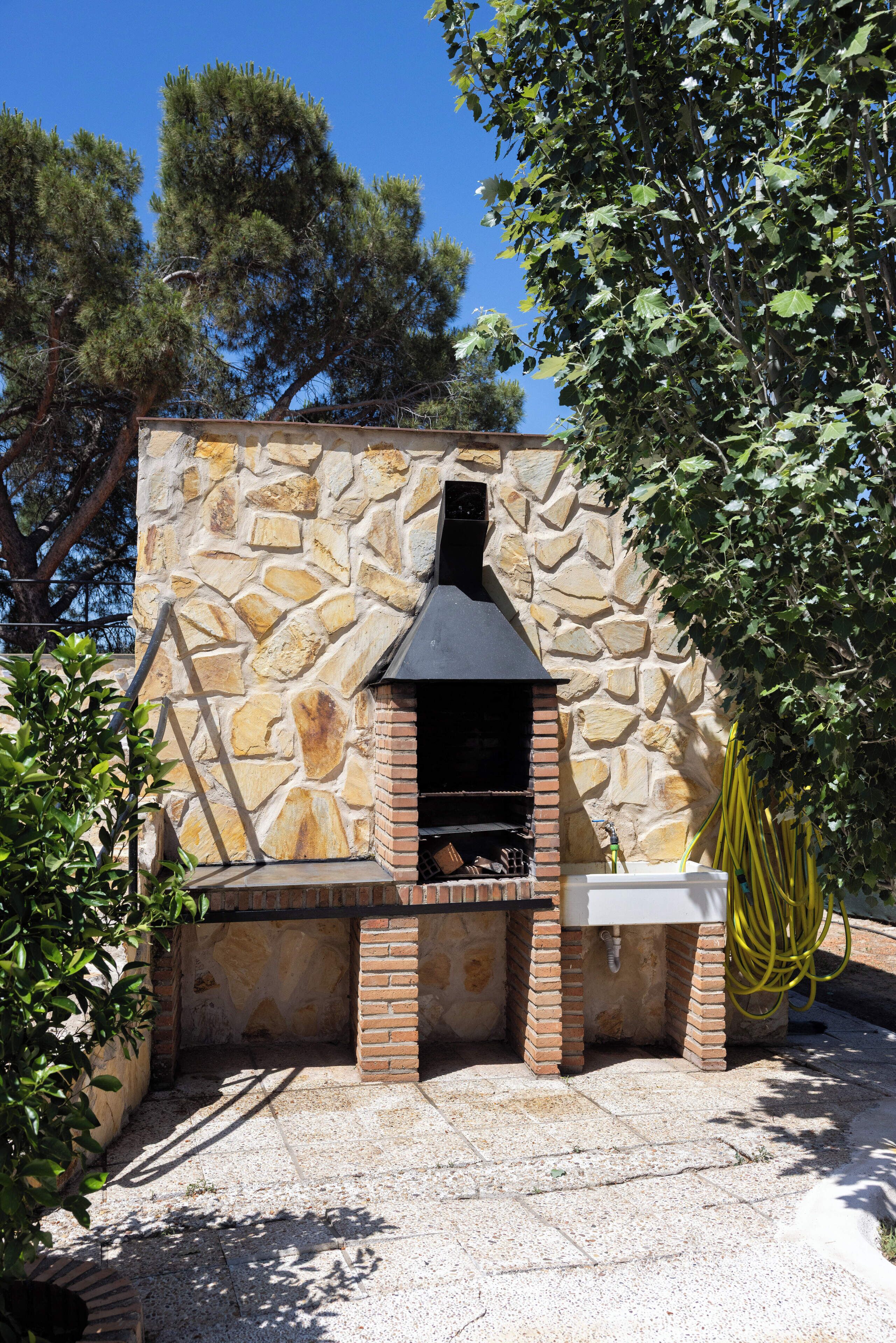 A stone-built outdoor kitchen with a chimney, surrounded by trees and a clear sky.