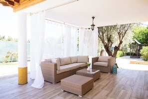 A covered outdoor seating area with a sofa and ottoman, a hanging light fixture, and a view of a garden and water feature.