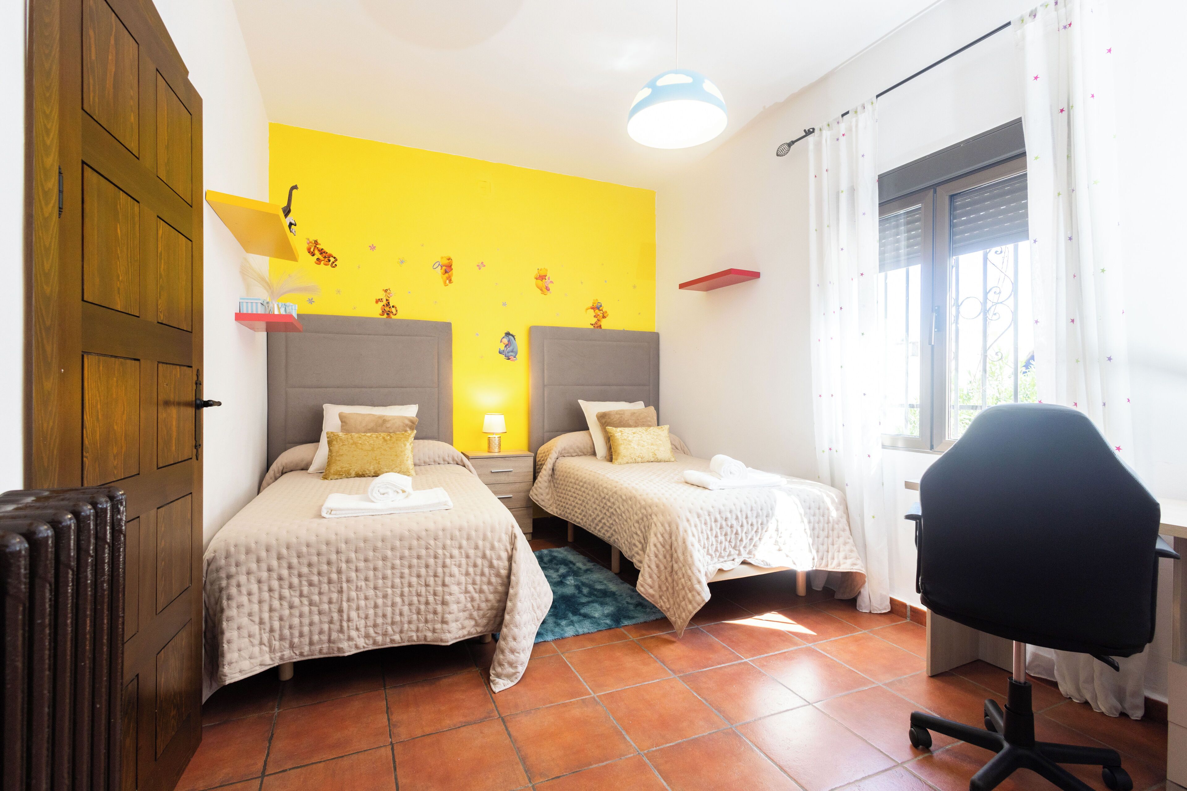 Una habitació amb dos llits, un escriptori, una cadira, un radiador i una finestra amb cortines.