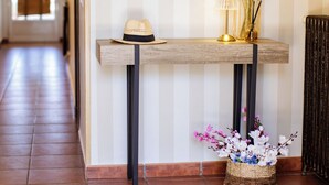 Un pasillo con pared a rayas, un espejo redondo, una consola de madera con un sombrero, una lámpara y un jarrón con flores.