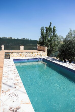 Une piscine rectangulaire à l’eau d’un bleu limpide, avec des bordures en pierre et en carrelage, et un mur de pierre sur lequel est adossée une clôture en métal, visible au fond.