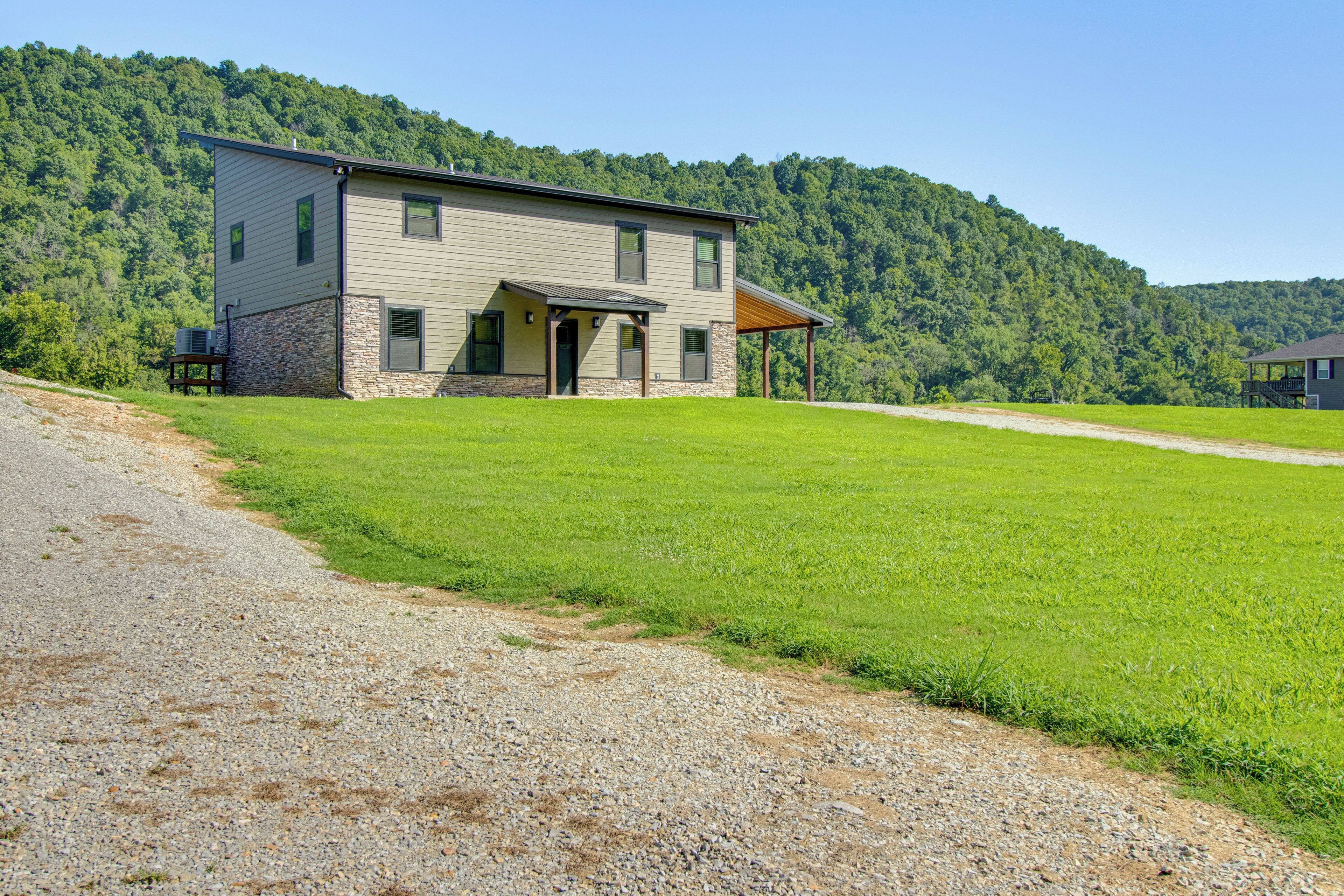 A two-story house with a covered porch, surrounded by a green field and a forested hillside.