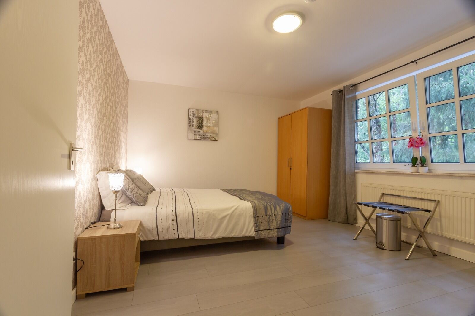 Un dormitorio con cama, mesita de noche, lámpara, armario, ventana con cortinas, radiador y una pequeña mesa con un cubo de basura.