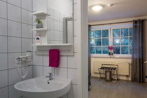 Un baño con un lavabo blanco, un secador de pelo, un estante con artículos de aseo y una ventana con vistas a árboles.