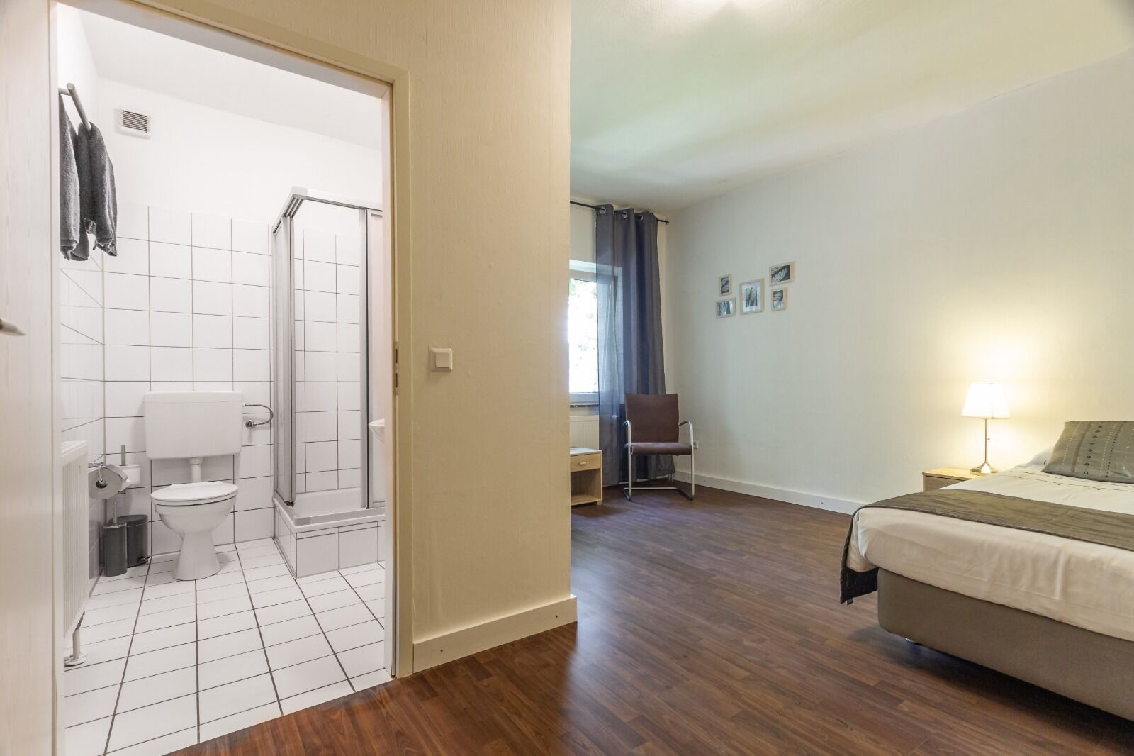 Een badkamer met toilet, douche en een stoel. De kamer heeft een houten vloer en een bed met een lamp.