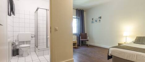 Een badkamer met toilet, douche en een stoel. De kamer heeft een houten vloer en een bed met een lamp.