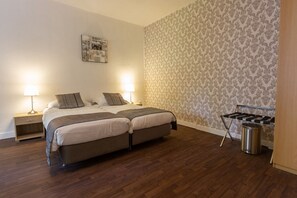 Habitación de hotel con dos camas, piso de madera, papel tapiz estampado y una plancha pequeña con un cubo de basura.