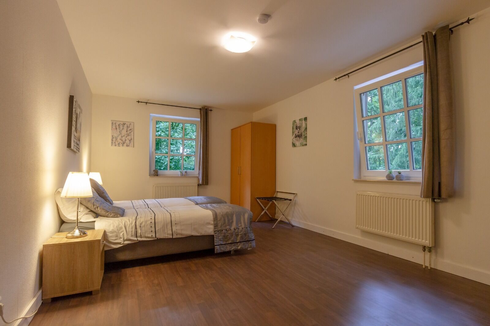 Un dormitorio con cama, mesita de noche, radiador y dos ventanas.