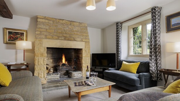 A living room with a stone fireplace, a television, and a sofa with yellow cushions. - Charming dog friendly cottage - Bank Cottage (Longborough)
