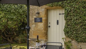 A table set outdoors with a bottle of wine, glasses, cheese, and bread. A sign reading "Bank Cottage" is visible on the wall. - Charming dog friendly cottage - Bank Cottage (Longborough)