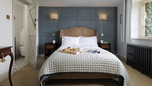 A neatly made bed with a floral bedspread, two bedside tables with lamps, a window with a view of greenery, and a radiator on the floor. - Charming dog friendly cottage - Bank Cottage (Longborough)