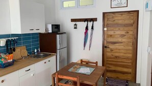 Cocina compacta con refrigerador, microondas y una mesa de comedor de madera con sillas. Hay un ventanal sobre el lavamanos, una puerta de madera y un perchero con paraguas.