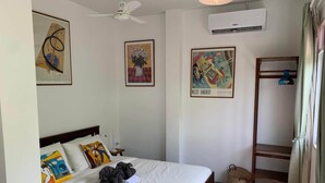 Habitación de hotel con una cama, ventilador de techo, equipo de aire acondicionado, armario y obras de arte en las paredes.