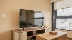 A modern living room with a flat-screen TV mounted on a wooden console, a small table with a decorative item, and a large window with blinds. - w Elegant 1BR Retreat in the Heart of San Isidro (Lima)