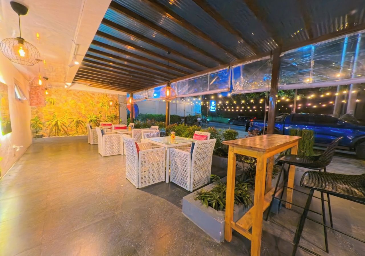 An outdoor seating area with wicker furniture and a glass table, surrounded by plants and string lights.