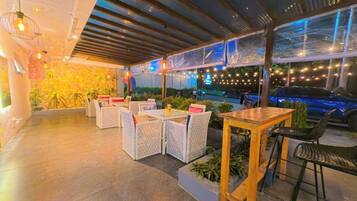 An outdoor seating area with wicker furniture and a glass table, surrounded by plants and string lights.