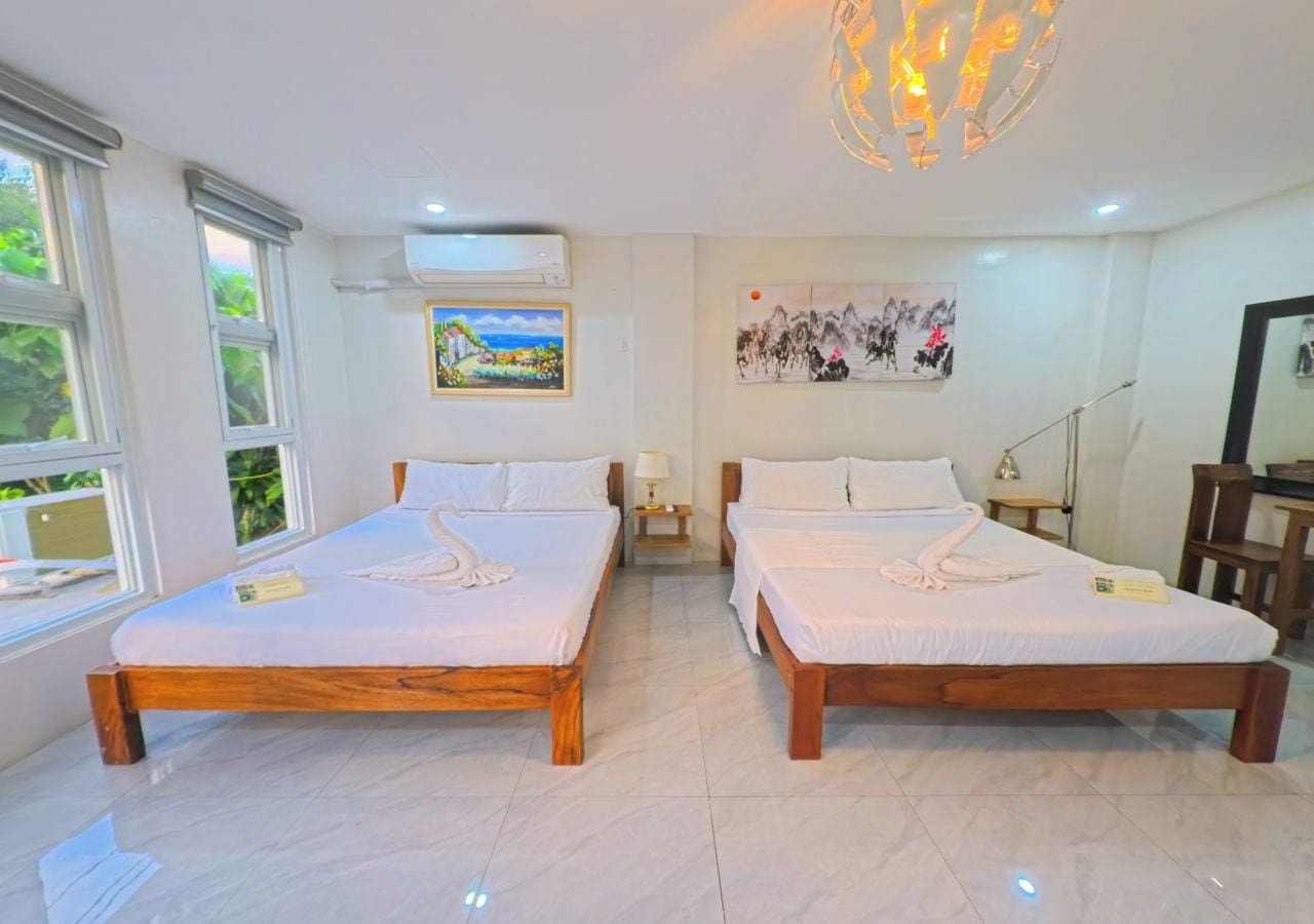 A hotel room with two wooden beds, a large window, an air conditioning unit, a painting, and a modern chandelier.