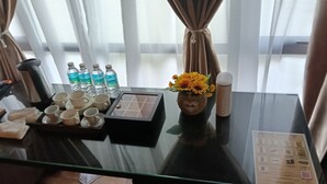 A hotel room with a glass table displaying a tray of cups, a vase with flowers, water bottles, and a remote control.