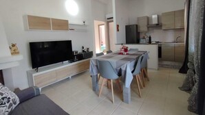A modern living room with a flat-screen TV, a dining table with chairs, and a kitchen area with cabinets and a refrigerator.