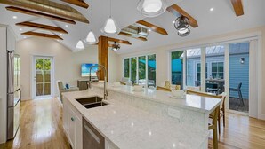 Een moderne keuken met een groot eiland, houten balken, hanglampen en uitzicht naar buiten.