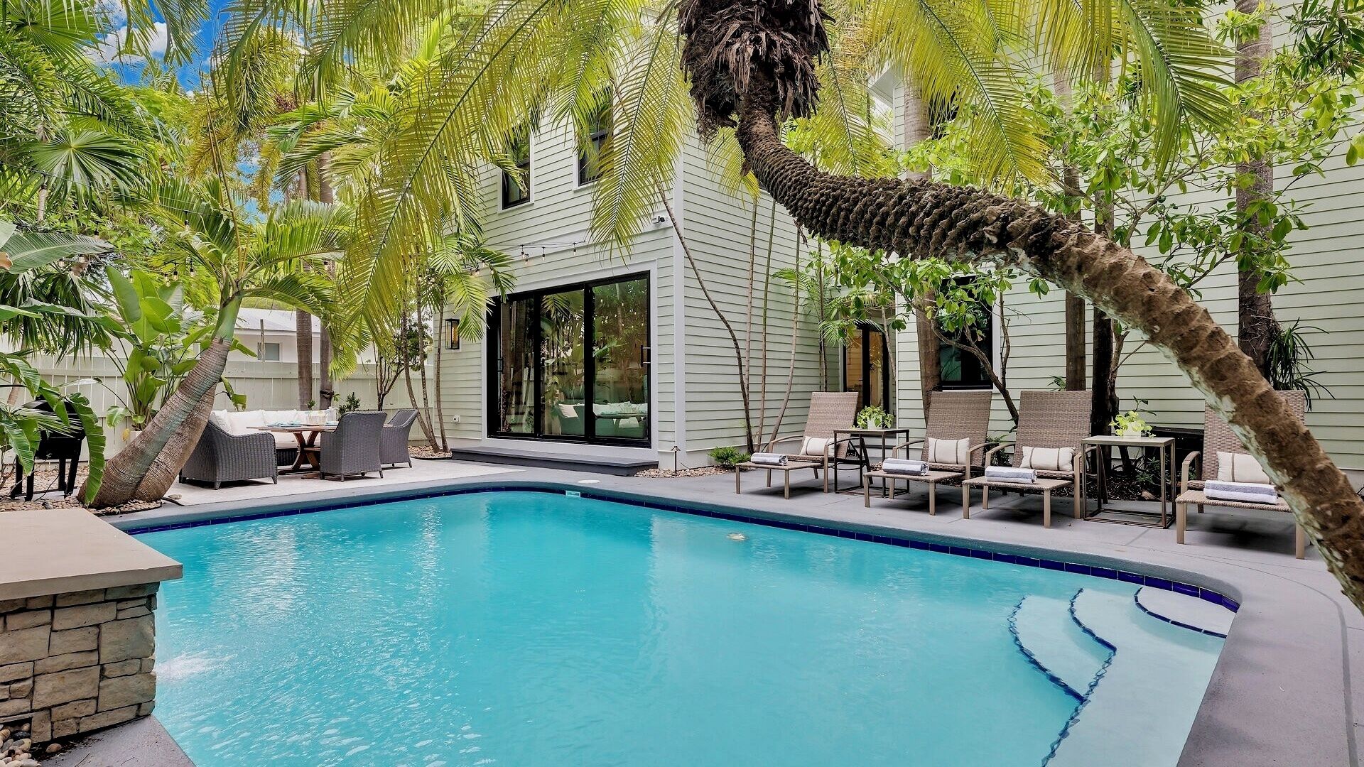 Une piscine entourée de chaises longues et d’un espace repas, le tout bordé de palmiers et adjacent à une maison dotée de grandes fenêtres.