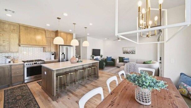 Une cuisine moderne avec des meubles en bois, un îlot central doté de tabourets, un éclairage suspendu et un coin repas avec une table et des chaises en bois.