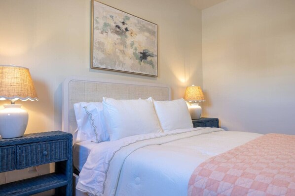 A neatly made bed with white linens and a pink quilt, flanked by two wicker nightstands with lamps, in a room with a plain wall and a single abstract painting. - Luxury 4BR |Sleeps 9 | New Build | Family Friendly (Broken Arrow)