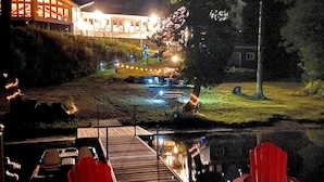 A wooden deck with red chairs, a dock extending into a body of water, and a building with illuminated windows in the background.